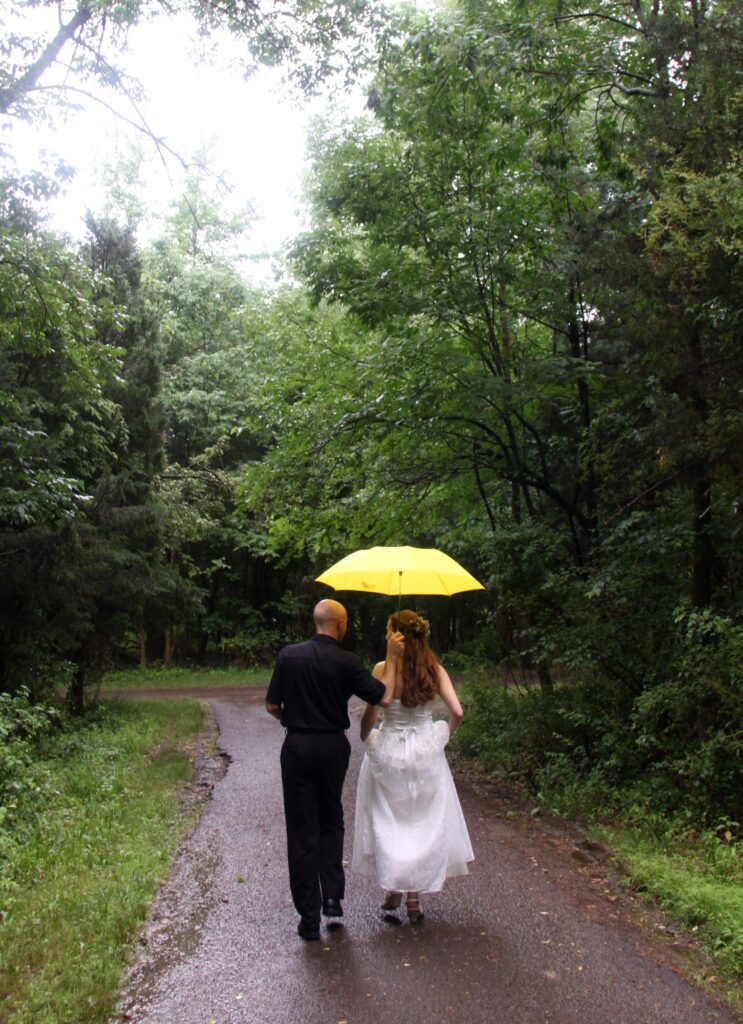 Photo taken from behind Jac and Angela on their wedding day as they walk down a wooded path under an umbrella