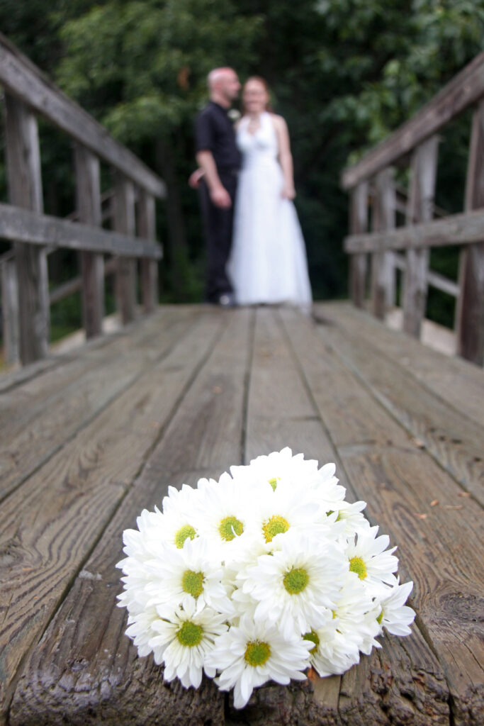 Wedding day photo of a bouquet of daisies in the foreground of a wooden pier, with Jac and Angela in the distant background.